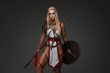 Beyaz manto, deri kayışlar, omuz koruyucuları, kürk ve baldırları olan, fantezi zırhlı kaslı sarışın bir Viking kadın, gri bir arka plana karşı kalkan ve mızrakla poz veriyor.