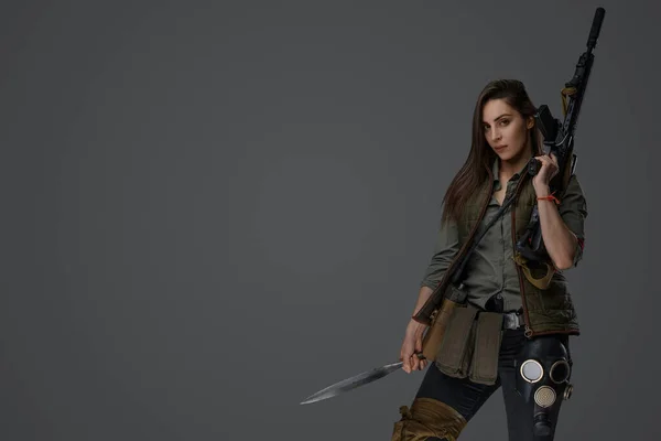 Middle Eastern woman, dressed in post-apocalyptic survivalist gear, poses with a dagger and an assault rifle against a gray background, embodying resilience and preparedness