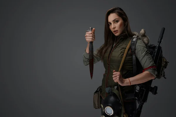 Middle Eastern descent in post-apocalyptic survival gear, brandishing a dagger on a gray backdrop, showcasing her determination to survive
