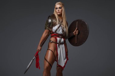 Çıplak bacaklı fantezi zırhlı Viking kadın. Gri arka planda kendine güvenen bir kalkanı ve mızrağı var.