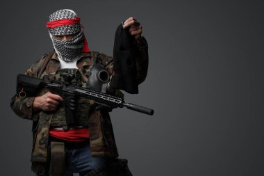 Orta Doğulu bir militan, beyaz keffiyeh giyip kamufle olmuş, elinde tüfekle gri bir fona karşı elinde duran siyah bir kar maskesi ile poz veriyor.