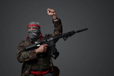 Orta Doğulu fanatik bir asker, beyaz keffiyeh giyiyordu ve kamufle olmuş bir üniforma giyiyordu, tüfekle silahlıydı, gri bir arka plana karşı elini kaldırmış poz veriyordu.