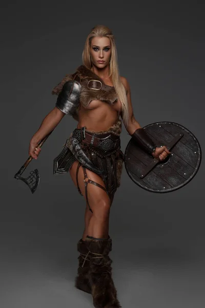 A fierce barbarian or Viking maiden with blonde hair wears lightweight armor, fur, and exposes her chest. She proudly holds an axe and shield against a gray background