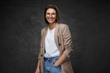 Neşeli, şık giyinmiş gözlüklü, tişörtlü ve ceketli bir kadın koyu bir arka plana karşı duruyor.