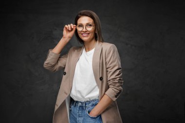 Neşeli, şık giyinmiş gözlüklü, tişörtlü ve ceketli bir kadın koyu bir arka plana karşı duruyor.