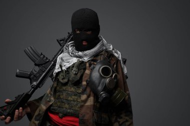 Orta Doğu 'dan radikal fanatik bir asker, siyah kar maskesi ve kamufle olmuş bir üniformayla gizlenmiş, otomatik tüfekli, gri bir arka planda.