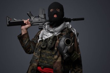 Siyah kar maskesi ve kamufle olmuş, otomatik tüfekle donatılmış, soğuk gri bir arka plana karşı silahlı bir Orta Doğulu fanatik askerin tehditkar portresi.