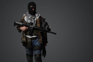 Orta Doğulu bir militanın portresi, siyah kar maskesi ve kamuflaj giyiyordu, elinde bir tüfek vardı, gri bir arkaplanda duruyordu.