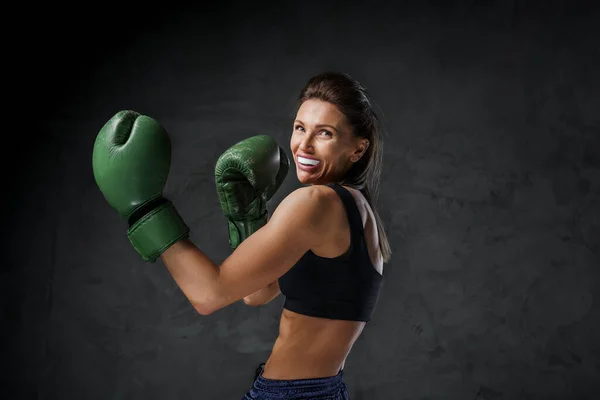 Athletic female fighter with a mouthguard, in sports bra, shorts, and boxing gloves, demonstrates striking skills on a dark background