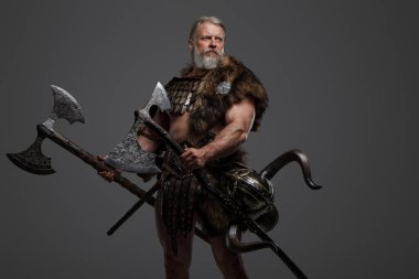 Kürk ve hafif zırh giymiş yaşlı sakallı Viking savaşçısı iki büyük balta tutuyor, gri bir arka plana karşı.
