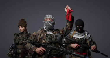 Üç Ortadoğulu militan grubu kamuflaj üniformaları, keffiyehler ve kar maskeleri giyip gri arka planda poz veriyorlar.
