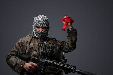 Orta Doğu radikal fanatik askeri, beyaz keffiyeh giymiş ve kamufle olmuş üniformalı, tüfekli, kırmızı bir bez ve uzaktan kumandayla elini kaldırmış.