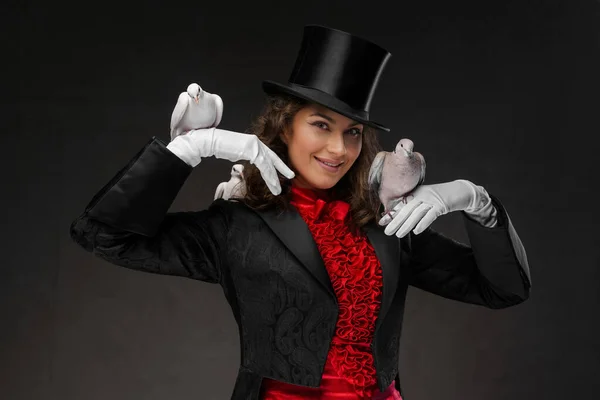 A portrait of a magician in a magicians costume and black top hat performing magic tricks with white doves against a dark background