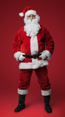 Geleneksel kırmızı takım elbiseli neşeli Noel Baba elleri kalçasında kırmızı arka planda duruyor.