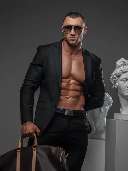 Rugged man in sunglasses, wearing an open suit jacket, showcasing his muscular torso, posing with a luxurious high-fashion bag next to ancient Greek statues on a gray background