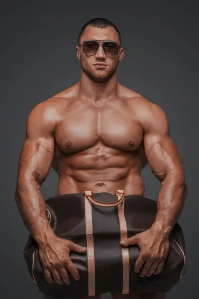頑丈な魅力 スポーツサングラスを除くハンサムな男は 彼の筋肉の胸を表示しながら 灰色の背景に絶妙なハイファッションバッグを保持しています — ストック写真