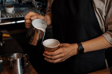 Arka planda bulanık bir espresso makinesi olan, kağıt bir kahve bardağına köpüklü süt döken bir baristanın yakın çekimi.