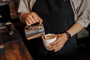 Arka planda bulanık bir espresso makinesi olan, kağıt bir kahve bardağına köpüklü süt döken bir baristanın yakın çekimi.
