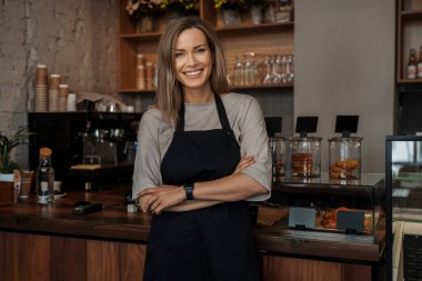 Önlüklü gülümseyen kadın rahat bir kafe ortamında güvenle duruyor, arka planda çiçek rafları var.