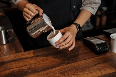 Arka planda bulanık bir espresso makinesi olan, kağıt bir kahve bardağına köpüklü süt döken bir baristanın yakın çekimi.