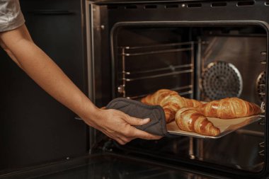 Bir insan, koruyucu bir fırın eldiveni takmış bir tepsi altın rengi kruvasanı fırından çıkarıyor.