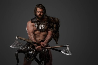 Kürk ve hafif zırhlı vahşi sakallı bir Viking savaşçısı, kemerine bağlı bir miğfer, gri arka planda büyük iki elli bir balta tutuyor.