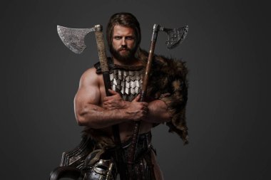 Kürk ve hafif zırh giymiş, kemerine kask takmış ve gri arka planda iki balta tutan haşin sakallı bir Viking savaşçısı.