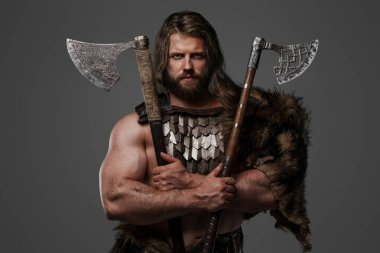 Kürk ve hafif zırh giymiş, kemerine takılı bir miğfer takmış, gri bir zemine karşı iki balta tutan heybetli sakallı bir Viking savaşçısı.