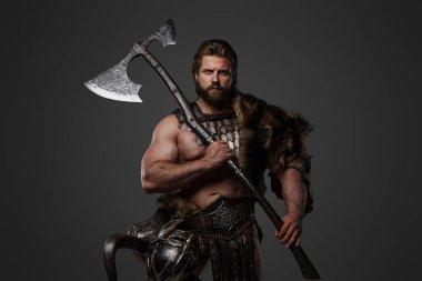 Kürk ve hafif zırhlı vahşi sakallı bir Viking savaşçısı, kemerine bağlı bir miğfer, gri arka planda büyük iki elli bir balta tutuyor.