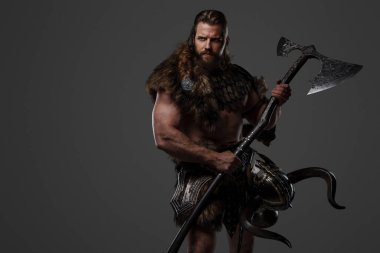 Kürk ve hafif zırhlı vahşi sakallı bir Viking savaşçısı, kemerine bağlı bir miğfer, gri arka planda büyük iki elli bir balta tutuyor.