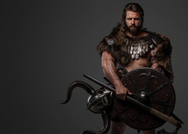 Kürk ve hafif zırh takmış, balta ve kalkan taşıyan, gri arka planda duran korkusuz bir Viking savaşçısı.