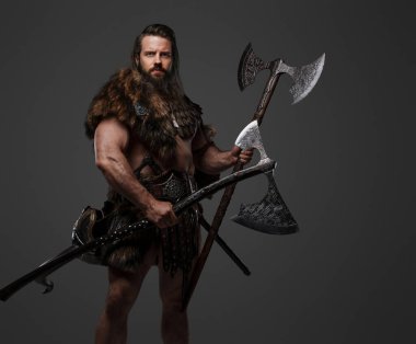 Kürk ve hafif zırh giymiş, kemerine kask takmış ve gri arka planda iki balta tutan haşin sakallı bir Viking savaşçısı.
