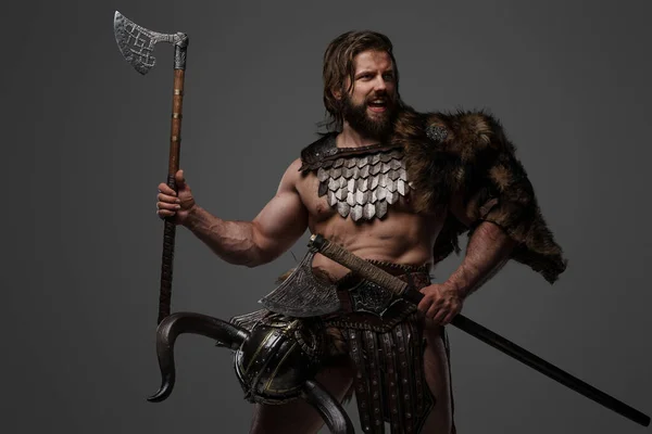 毛皮と軽装甲の激しいヴァイキングの戦士は 彼のベルトに取り付けられたヘルメットで 灰色の背景に大きな2手の斧を保持しています — ストック写真