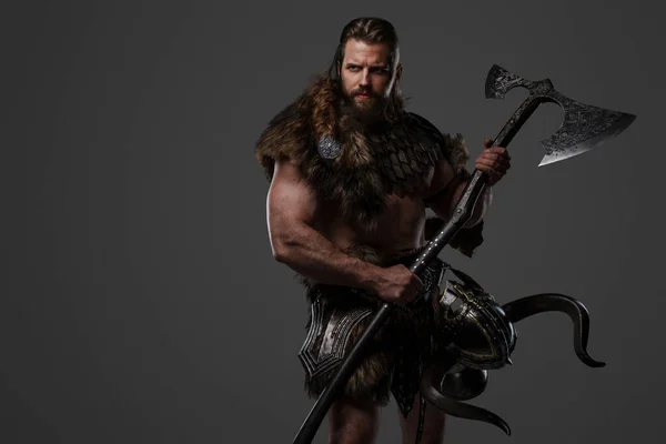 A fierce bearded Viking warrior in fur and light armor, with a helmet attached to his belt, holding a large two-handed axe on a gray background