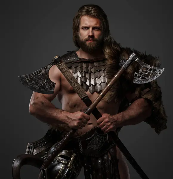 A rugged bearded Viking warrior dressed in fur and lightweight armor, with a helmet attached to his belt and holding two axes against a gray background