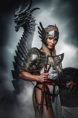 Kanatlı zırhlı yiğit Valkyrie arkasında bir ejderha ile nöbet tutuyor, karamsar, bulutlarla dolu bir gökyüzüne karşı.