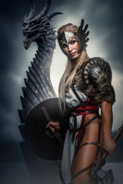 Kanatlı zırhlı yiğit Valkyrie arkasında bir ejderha ile nöbet tutuyor, karamsar, bulutlarla dolu bir gökyüzüne karşı.