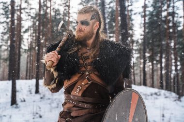 Karlı bir çam ormanında baltası ve kalkanı olan vahşi bir Viking savaşçısı, gücünü ve tarihsel İskandinav kültürünü tasvir ediyor.