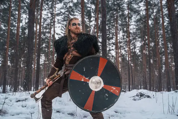 バイキングの戦士として服を着た男は 雪の森で斧と盾で立って 歴史的なノルウェーのイメージを呼び起こします — ストック写真