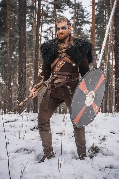 A man dressed as a Viking warrior stands with an axe and shield in a snowy forest, evoking historical Norse imagery