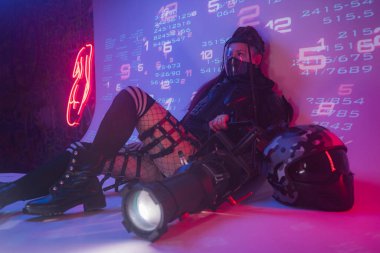 Fütüristik taktiksel siyah takım elbiseli bir kadın motosiklet kaskı ve büyük bir projektörle yerde yatıyor.