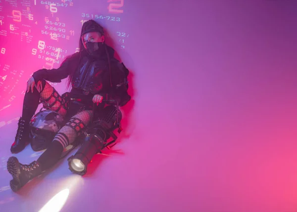 一位身穿未来派战术黑色西服的女人躺在地板上 身边躺着一个摩托车头盔和一个巨大的投影仪 背对着投射出来的数字符号 — 图库照片