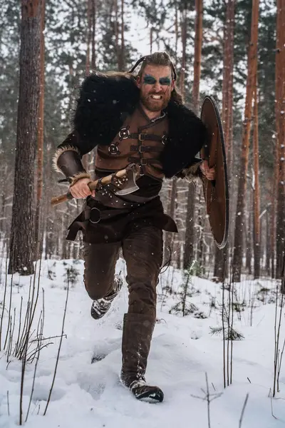 密集した森で雪が降る中で斧と盾で充電するバイキング戦士のダイナミックなイメージ — ストック写真