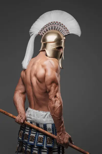 An impressive portrait of a bare-chested ancient Greek hoplite, wearing a helmet and brandishing a spear, standing in a studio with a gray backdrop