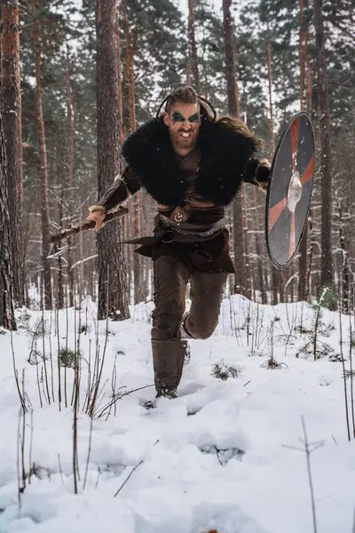 在茂密的森林里 一个维京战士拿着斧头和盾牌冲向雪花的动态画面 — 图库照片