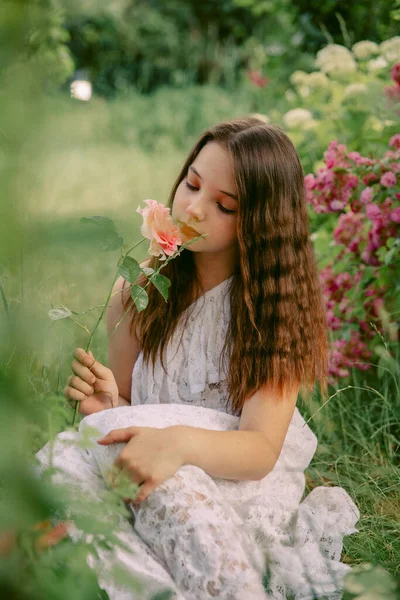 一个少女 手里拿着一朵玫瑰花 坐在盛开的花园里的草地上 — 图库照片