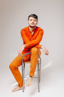 Parlak turuncu elbiseli bir adam bacağı havada bir sandalyeye oturur ve kamera lensine bakar.