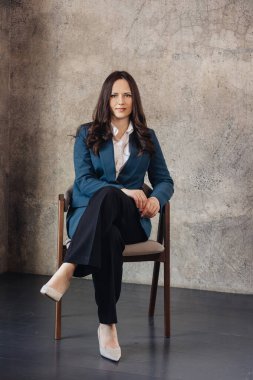 İş elbisesi giymiş genç bir kadın sandalyeye oturmuş, kendine güvenerek kameraya bakıyor. İş, profesyonellik ve kurumsal kavramlar için ideal..