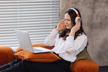 Kulaklık takan bir kadın elinde dizüstü bilgisayarla koltukta oturup uzaktan çalışmaktan zevk alıyor. Modern dijital iş ve internet iletişimini temsil eden meşgul ve profesyonel birine benziyor..