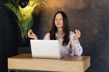 Bir iş kadını masasında dizüstü bilgisayarla meditasyon yapıyor, iş yerinde denge ve rahatlama arıyor..